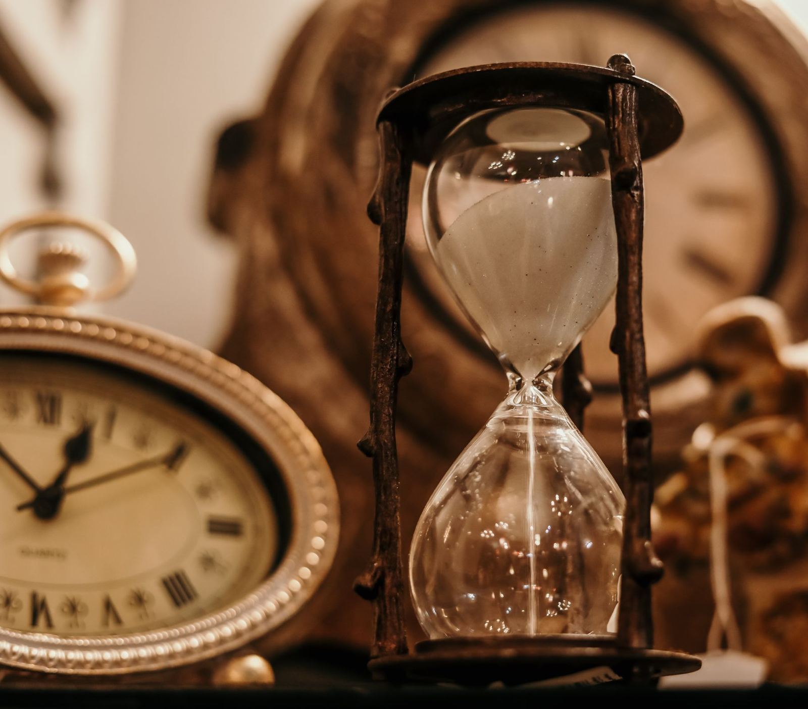 An hourglass with sand running through, beside two old clocks
