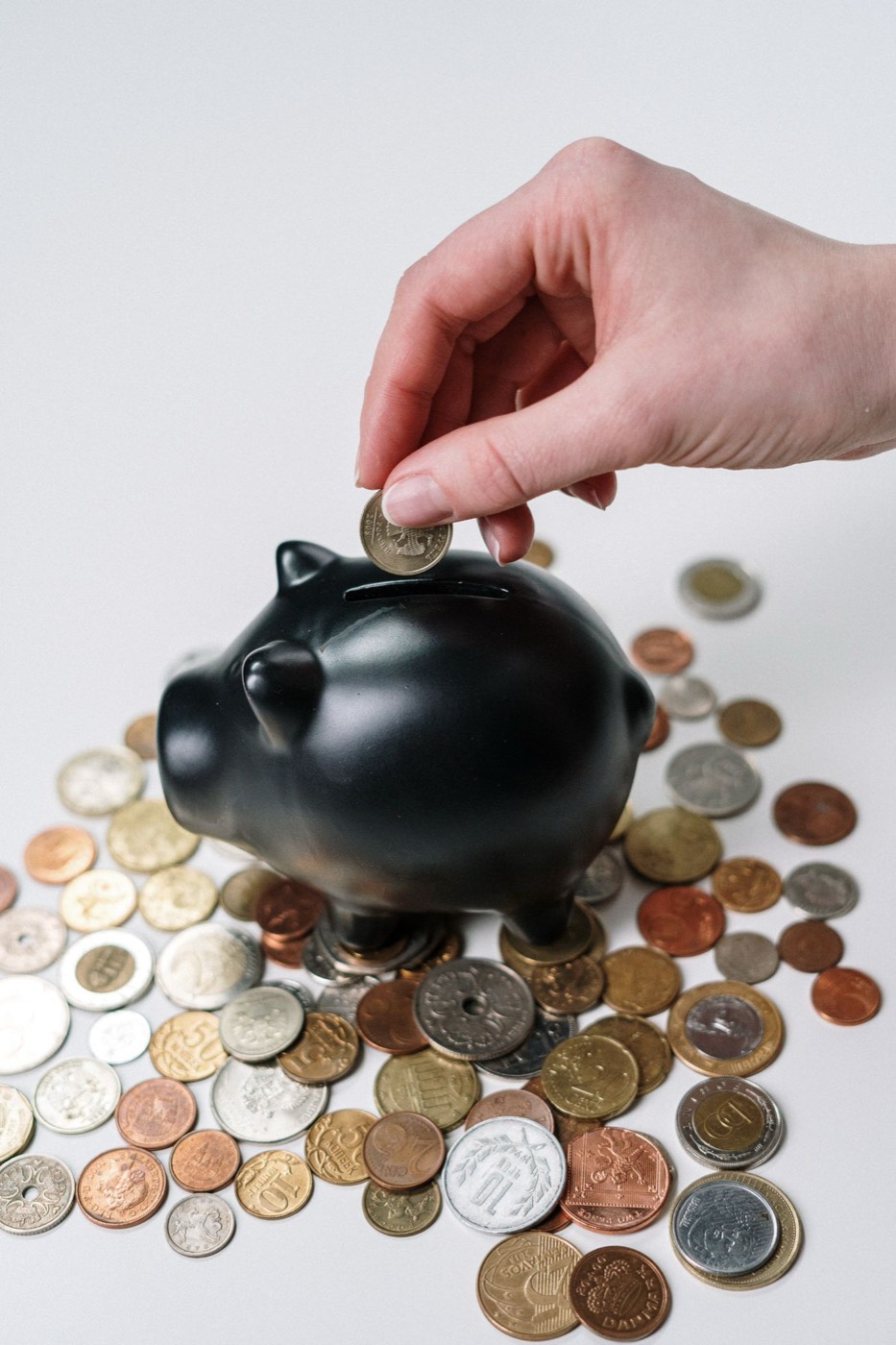 Hand dropping a coin into a black piggy bank surrounded by coins